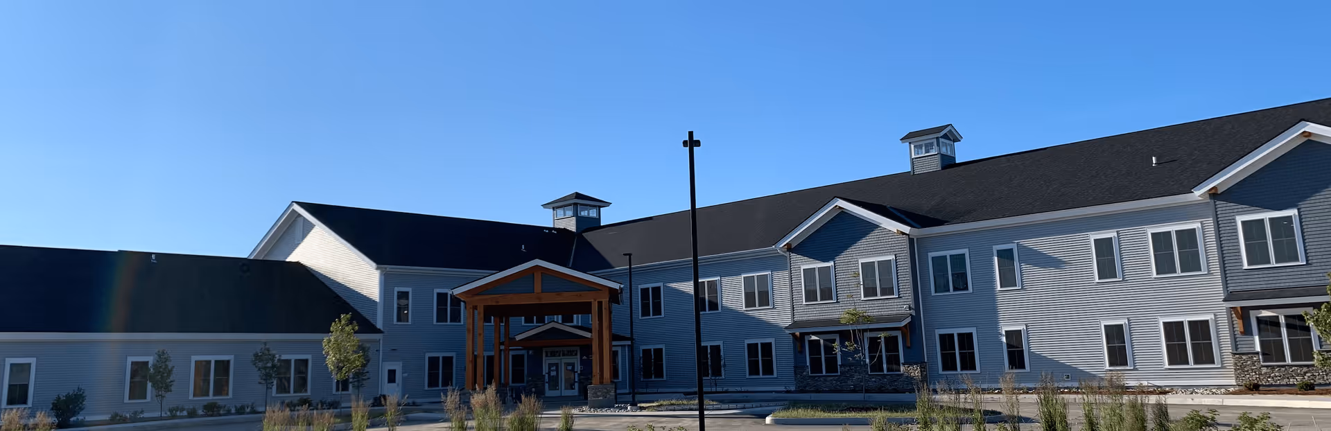 Exterior view of Maple Ridge Lodge Assisted Living facility showing a large two-story building with gray siding, multiple windows, and a covered entrance with wooden beams under a clear blue sky.