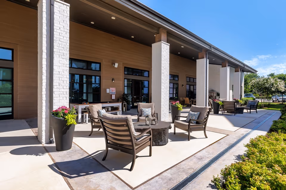 Outdoor patio area at Album Benbrook with cushioned chairs arranged around a circular table, large potted plants with flowers, and a covered walkway supported by white brick columns under a clear blue sky.
