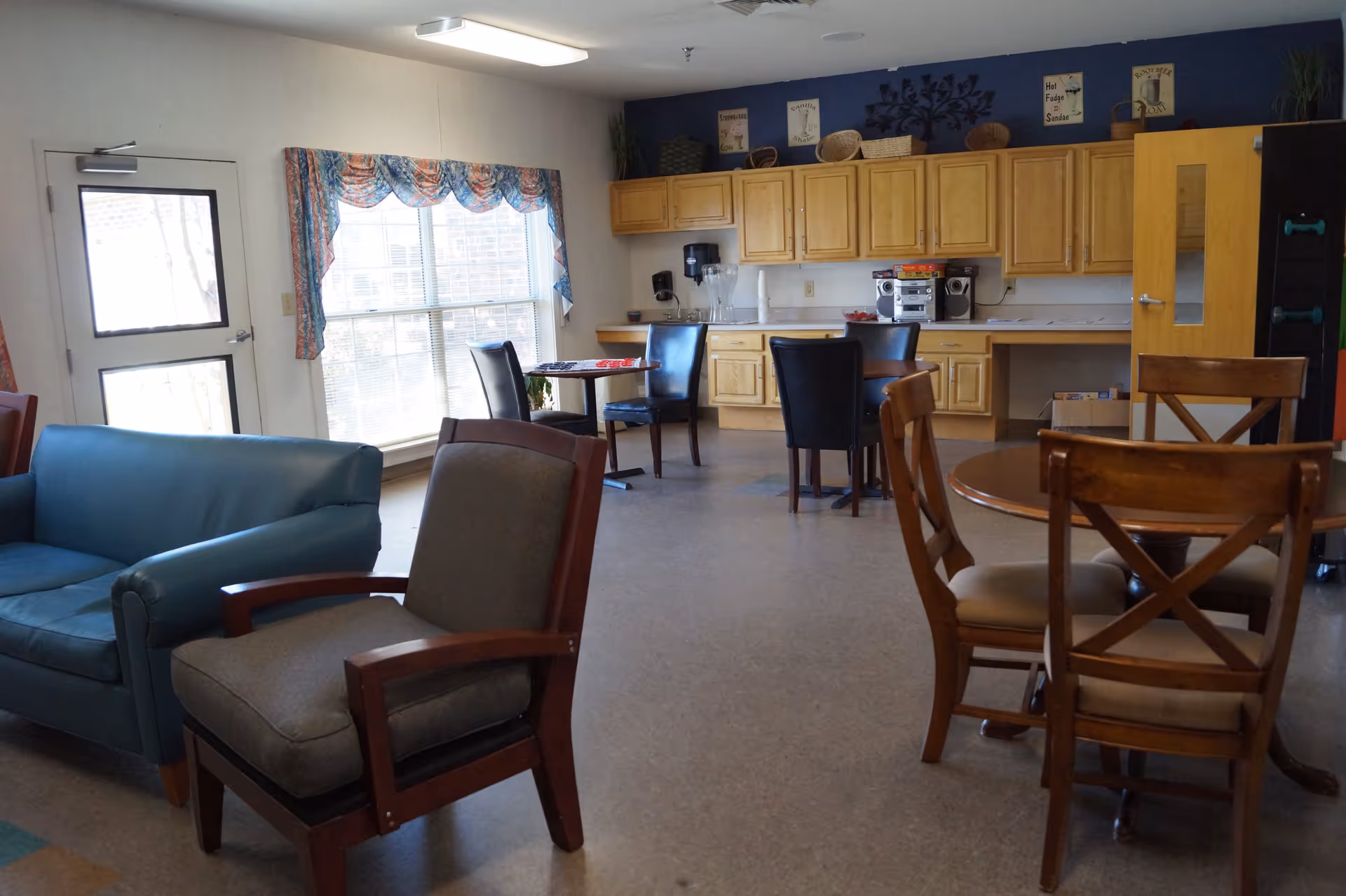 A common area in Zebulon House featuring a blue couch, a gray armchair, wooden chairs around a round table, and two small tables with black chairs. The back wall has wooden cabinets, a countertop with a stereo system, a water dispenser, and decorative signs. A large window with colorful curtains lets in natural light.