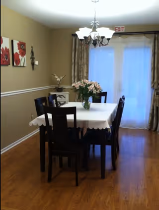 A dining room with a rectangular table covered with a white tablecloth and six dark wooden chairs around it. A vase with pink flowers is placed in the center of the table. The room has wooden flooring, beige walls with white trim, and three red floral paintings on the left wall. A chandelier with multiple lights hangs above the table, and there are large windows or glass doors with sheer curtains and heavier drapes in the background. An exit sign is visible above the windows.