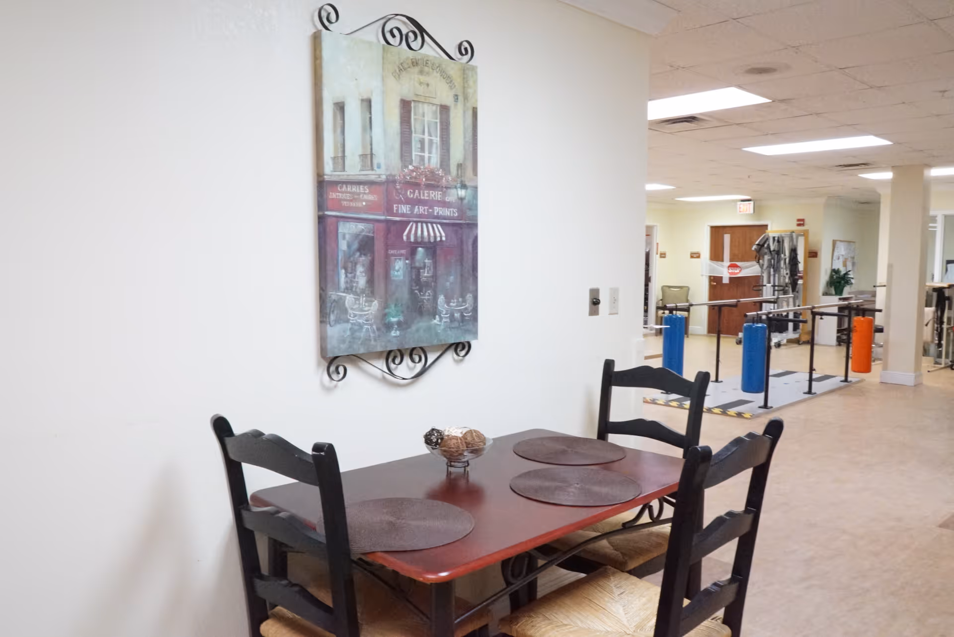 A small dining area with a wooden table and four chairs, each with a round placemat. On the wall above the table is a decorative painting of a storefront with the text 'Galerie Fine Art - Prints'. In the background, there is an open space with exercise equipment and parallel bars, suggesting a rehabilitation or physical therapy area.