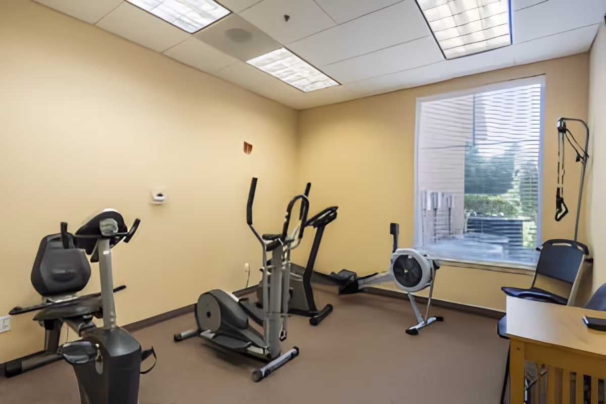 Small exercise room with stationary bikes, an elliptical, a rowing machine, and chairs by a window.