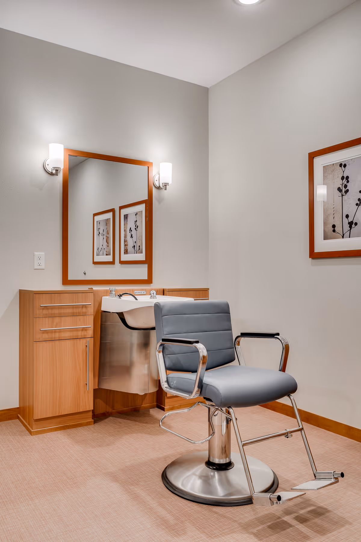 A modern salon area with a gray salon chair in front of a wooden cabinet and a sink. A large mirror with two wall-mounted lights is above the sink, and framed artwork is visible on the walls.