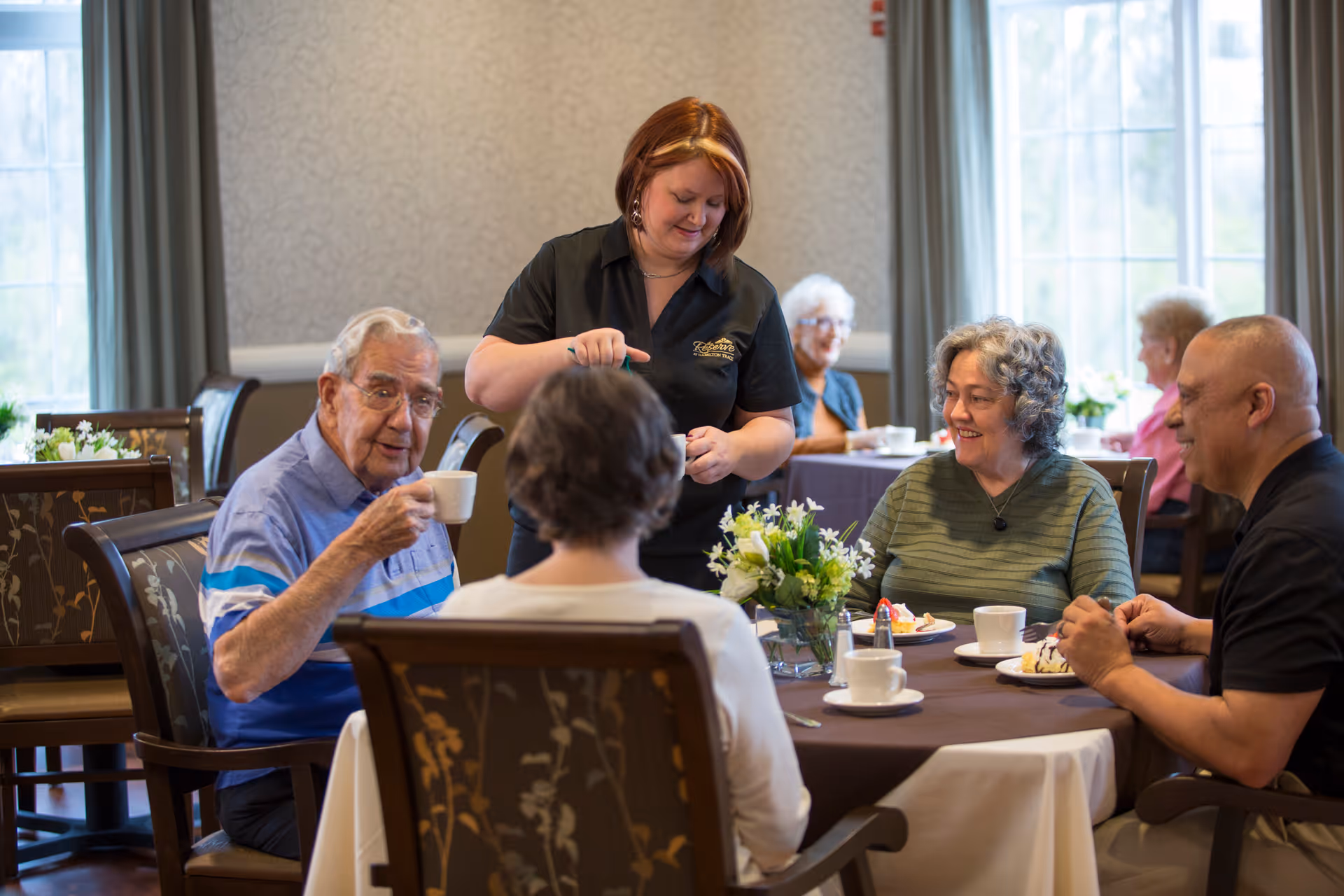 A staff member pours coffee for a group of seniors seated around a table in a communal dining room.