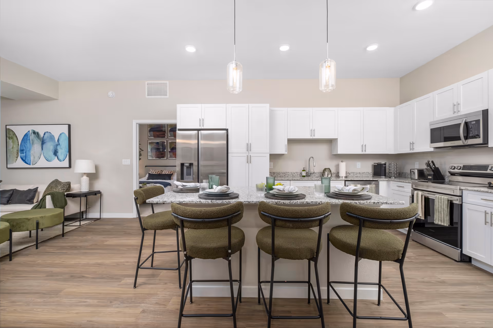 Modern kitchen with white cabinets, stainless steel appliances, and a large granite island with four green cushioned bar stools. The kitchen is well-lit with recessed ceiling lights and two pendant lights hanging over the island. To the left, part of a living area with a sofa, side table, lamp, and abstract wall art is visible. A doorway leads to a bedroom in the background.