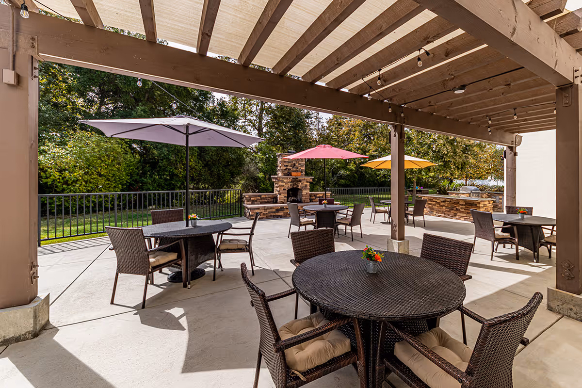 Outdoor patio area with several round wicker tables and cushioned chairs under a wooden pergola. Each table has a small flower arrangement and some tables have large colorful umbrellas. In the background, there is a stone fireplace and a built-in grill area surrounded by greenery and trees.