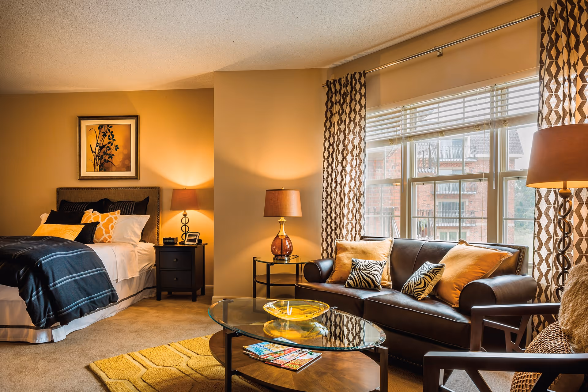 A cozy living space featuring a bed with black and yellow pillows and a dark blanket, a nightstand with a lamp and clock, a brown leather sofa with patterned cushions, a glass coffee table with magazines and a decorative bowl, and large windows with patterned curtains letting in natural light.
