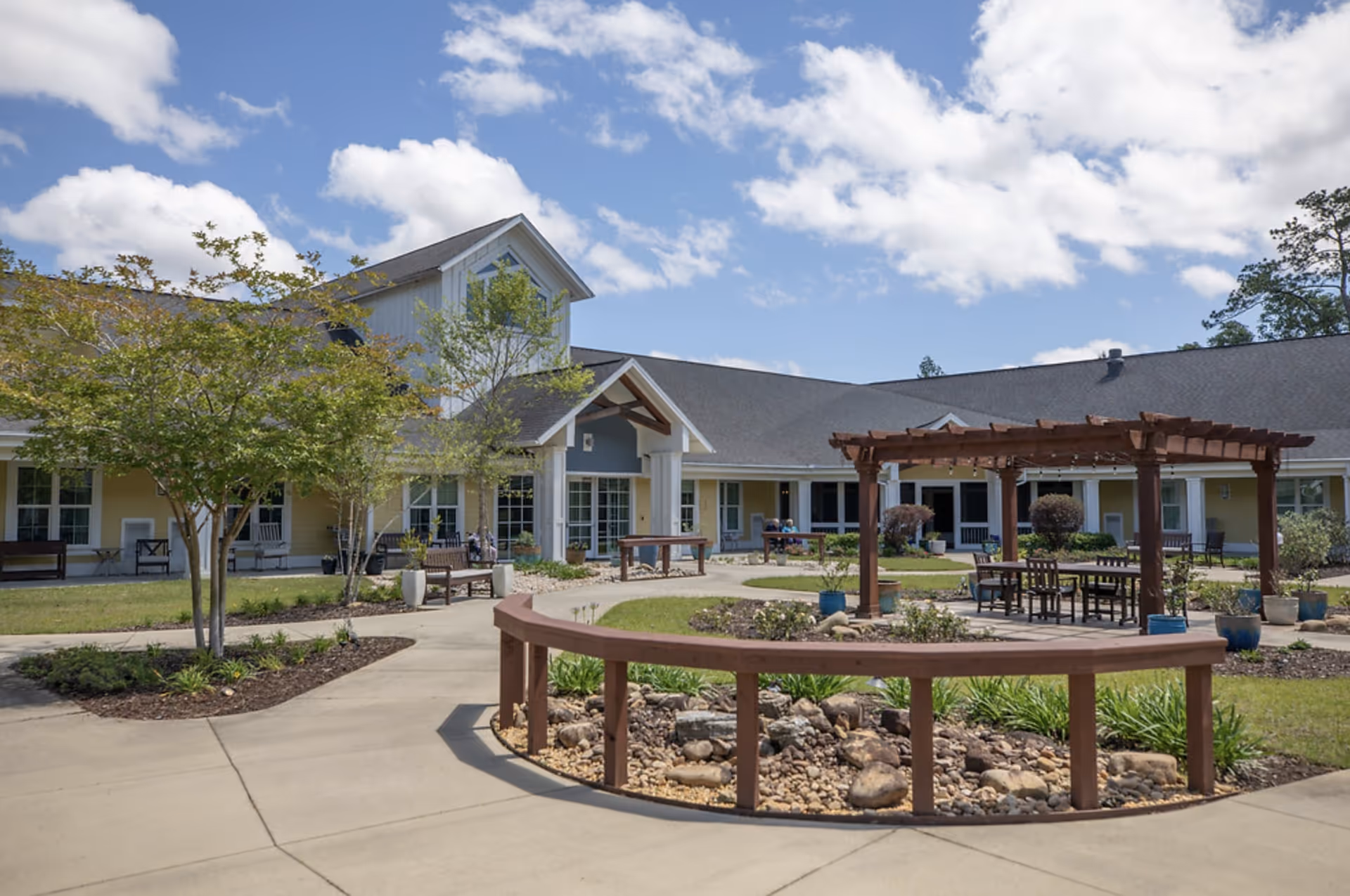 Outdoor courtyard area of a senior living facility with a circular garden bed filled with rocks and plants, a wooden pergola with seating underneath, benches along the walkway, and a building with multiple windows and a peaked roof in the background under a partly cloudy blue sky.