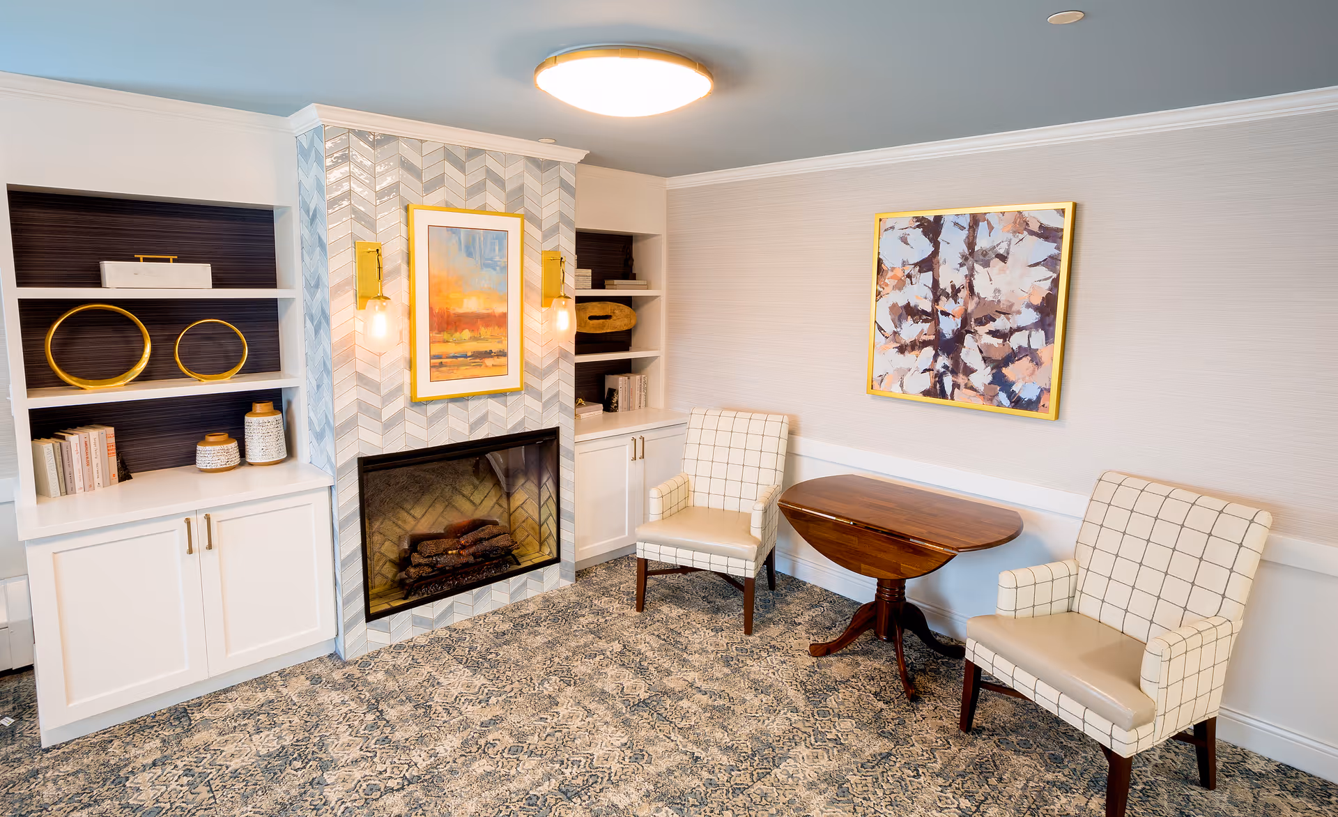 A cozy sitting area with two checkered armchairs and a wooden table between them. There is a fireplace with a herringbone tile surround and two wall sconces with exposed bulbs. Above the fireplace is a framed abstract painting with warm colors. Built-in white shelves with decorative items and books flank the fireplace. Another abstract painting with a gold frame hangs on the adjacent wall. The floor is covered with a patterned carpet.