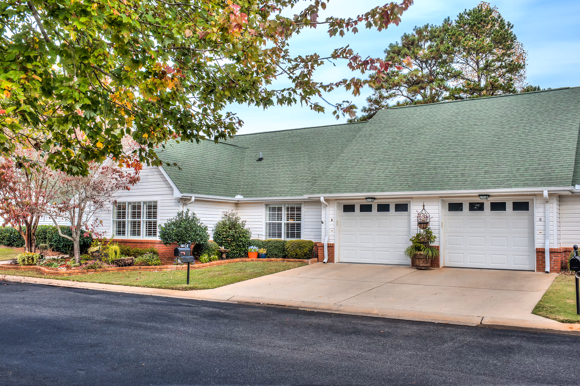 Exterior view of a single-story residential building with a green roof, white siding, and two white garage doors. The front yard is landscaped with bushes, small trees, and a mailbox near the driveway. A tree with green and reddish leaves partially frames the top left of the image.
