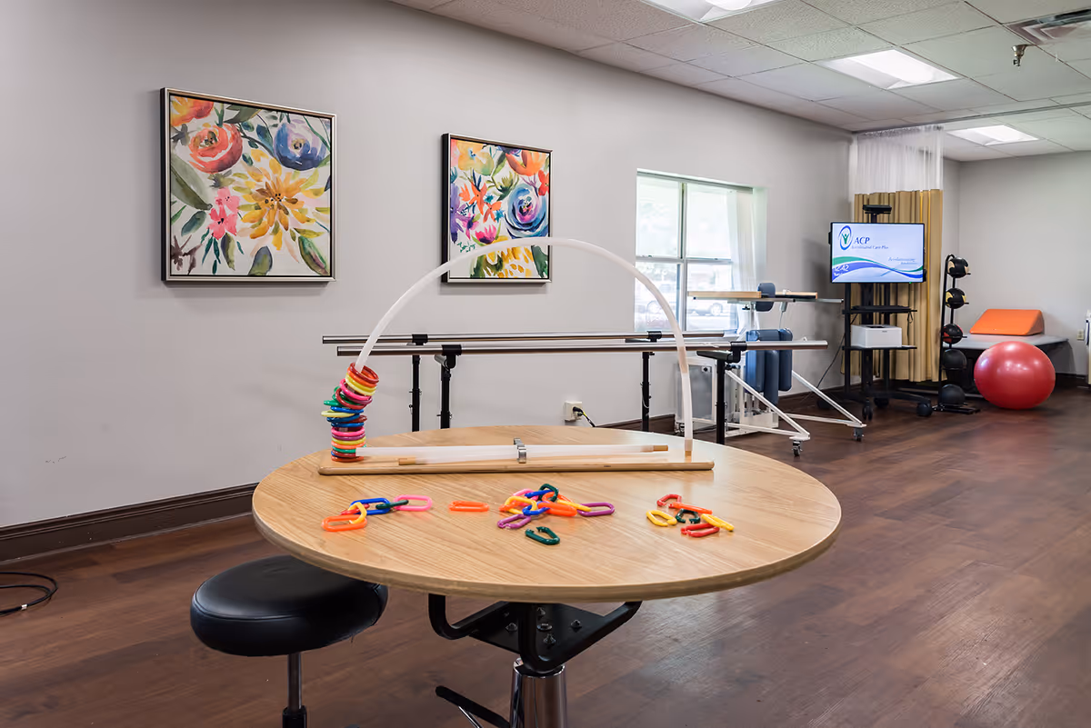 Activity/therapy room with a round table holding colorful plastic links and a ring-stacking toy, parallel bars, exercise equipment, and a TV on a cart.