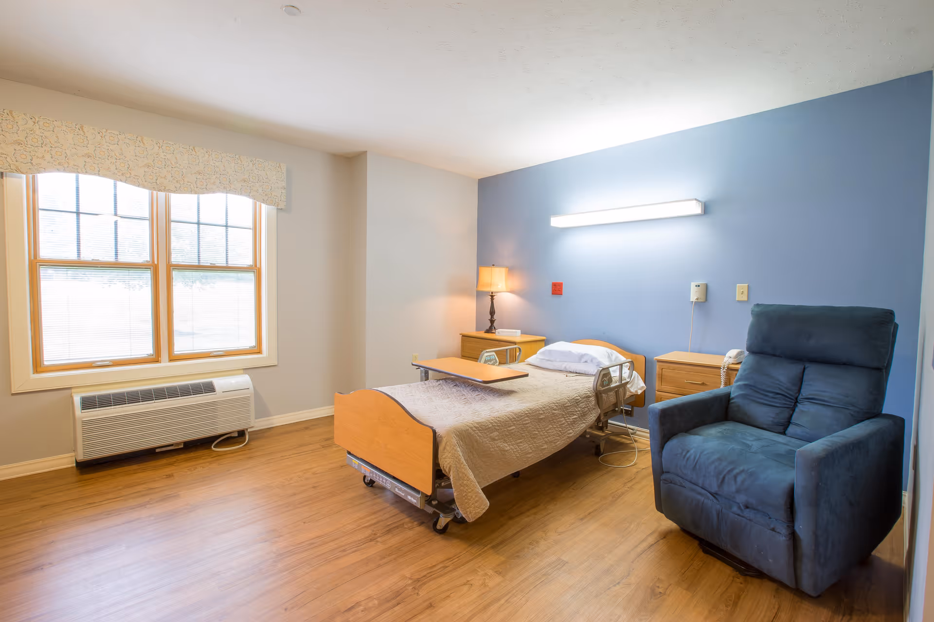 A bright resident room with a hospital-style bed, overbed table, nightstands and lamp, a blue accent wall, and a blue recliner beside a large window.