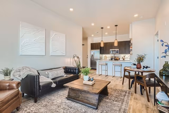A modern living room and kitchen area in an apartment. The living room features a black leather sofa with cushions and a throw blanket, a brown leather armchair, a wooden coffee table with a plant and books, and a patterned rug. The kitchen has dark cabinets, stainless steel appliances including a refrigerator and microwave, a white countertop island with two bar stools, and pendant lights hanging above. A small round dining table with two chairs and a vase with flowers is also visible. The walls are light-colored with two abstract white wall art pieces above the sofa.