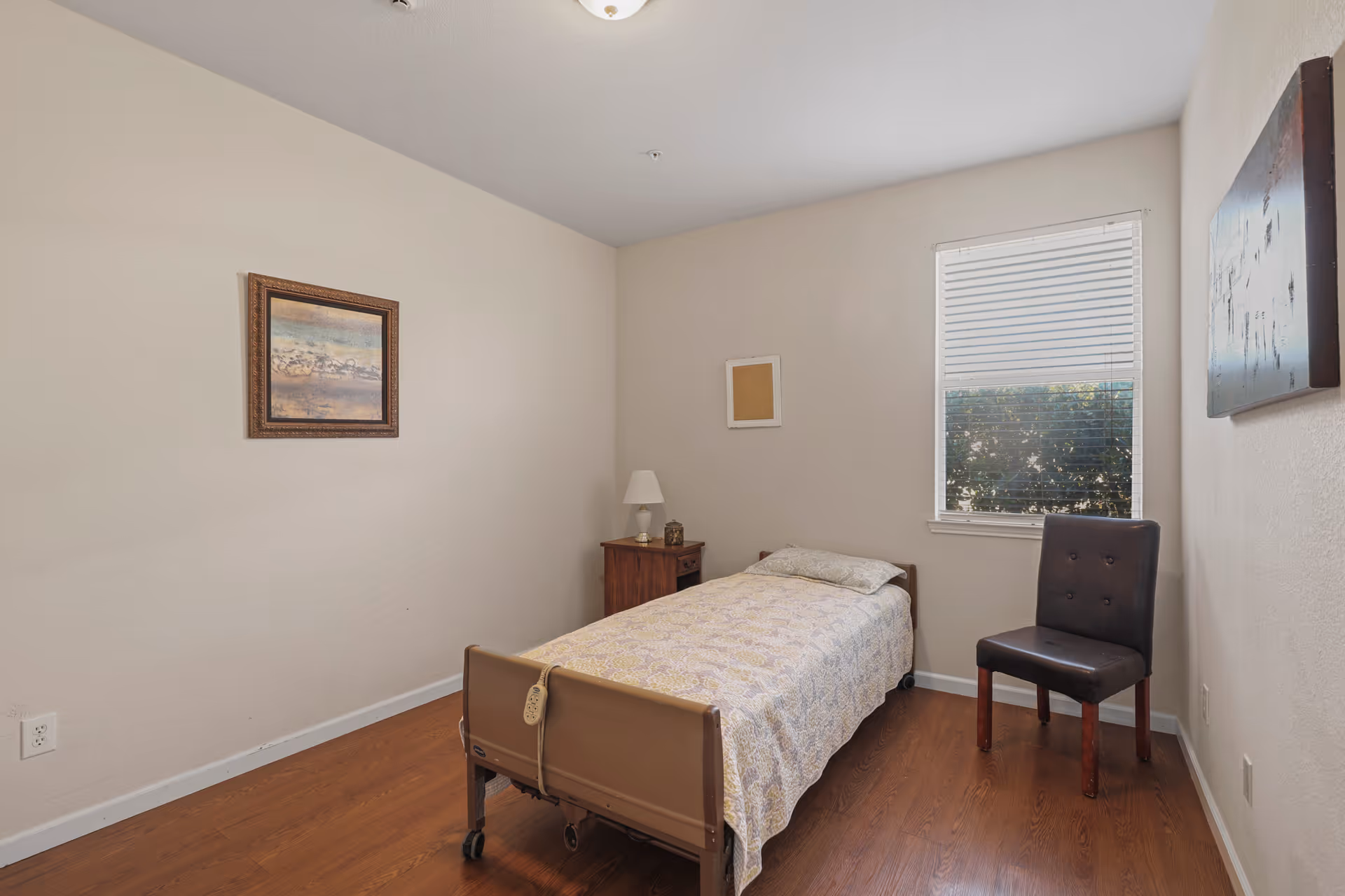A simple senior living bedroom with a single bed covered with a patterned bedspread, a wooden nightstand with a lamp, a dark cushioned chair, two framed pictures on the walls, and a window with blinds partially open showing greenery outside.