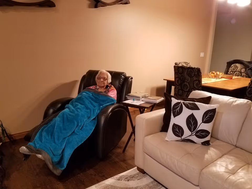 A person reclining in a black leather chair covered with a blue blanket in a living room beside a beige sofa and dining table.