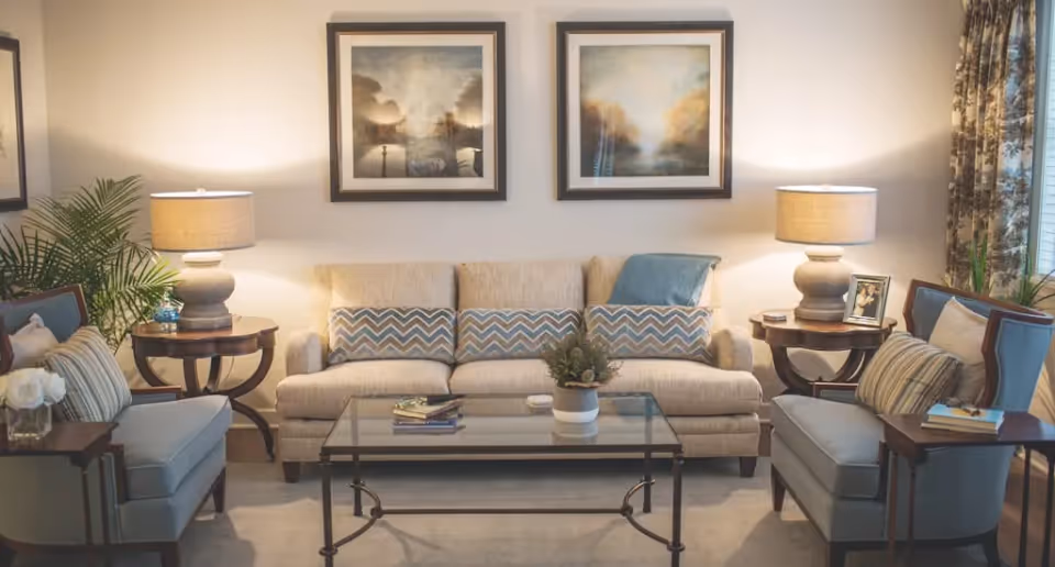 A cozy living room with a beige sofa adorned with patterned cushions and a blue throw blanket. Two matching armchairs with striped pillows flank a glass coffee table with books and a small plant. Two table lamps on round wooden side tables provide warm lighting. Two framed landscape paintings hang on the wall above the sofa. There are plants and floral curtains near a window on the right side.