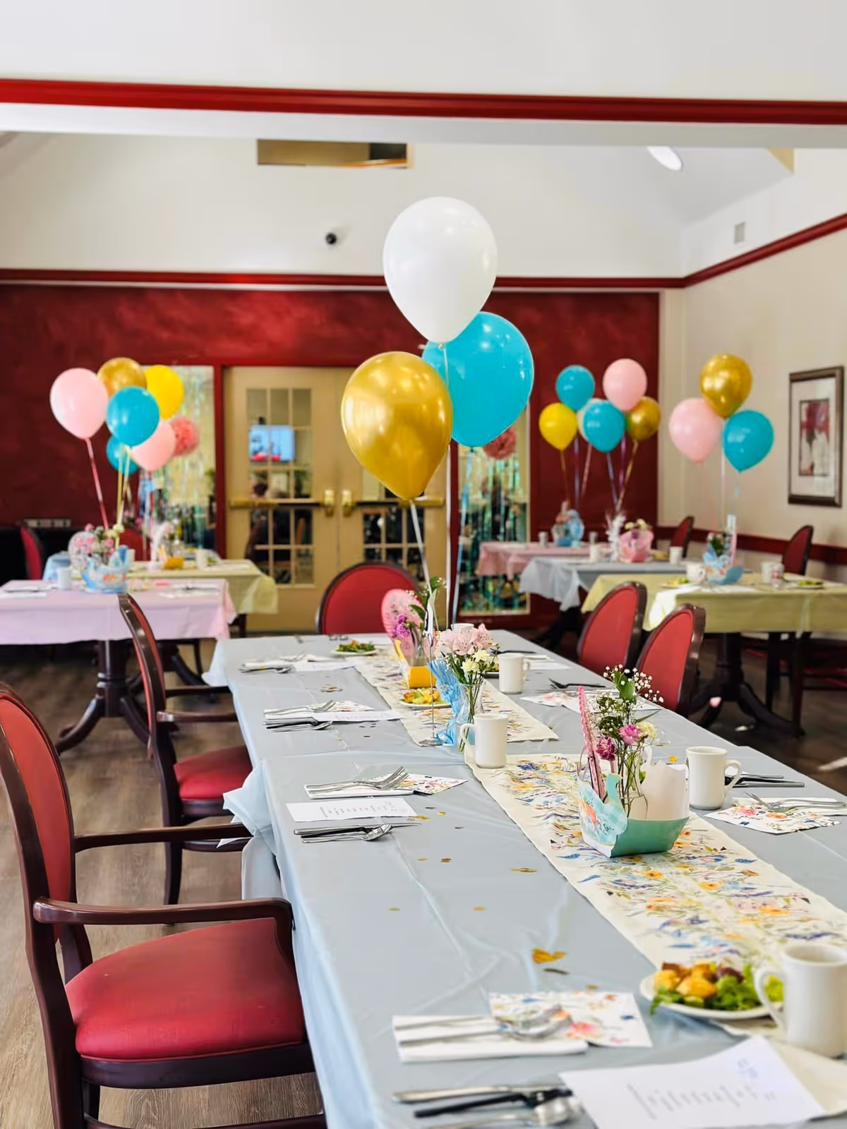 A decorated dining room with tables covered in pastel-colored tablecloths and floral runners. Each table is set with plates, cups, utensils, and small flower arrangements. Colorful balloons in white, gold, teal, pink, and yellow are tied to the chairs and centerpieces. The room has red walls with white trim and wooden flooring, with double doors and framed artwork on the walls.