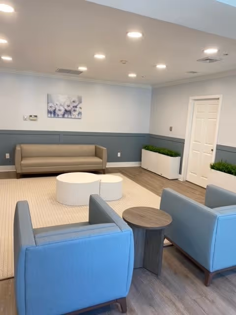 Bright communal lounge with a sofa, two blue armchairs, round coffee tables, and planters against a light-blue wainscoted wall.