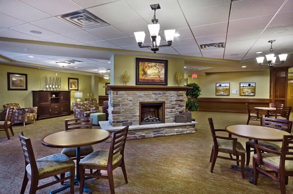 A spacious senior living common area with round wooden tables and chairs, a stone fireplace in the center, comfortable armchairs with patterned upholstery, framed landscape paintings on the walls, and ceiling lights providing warm illumination.