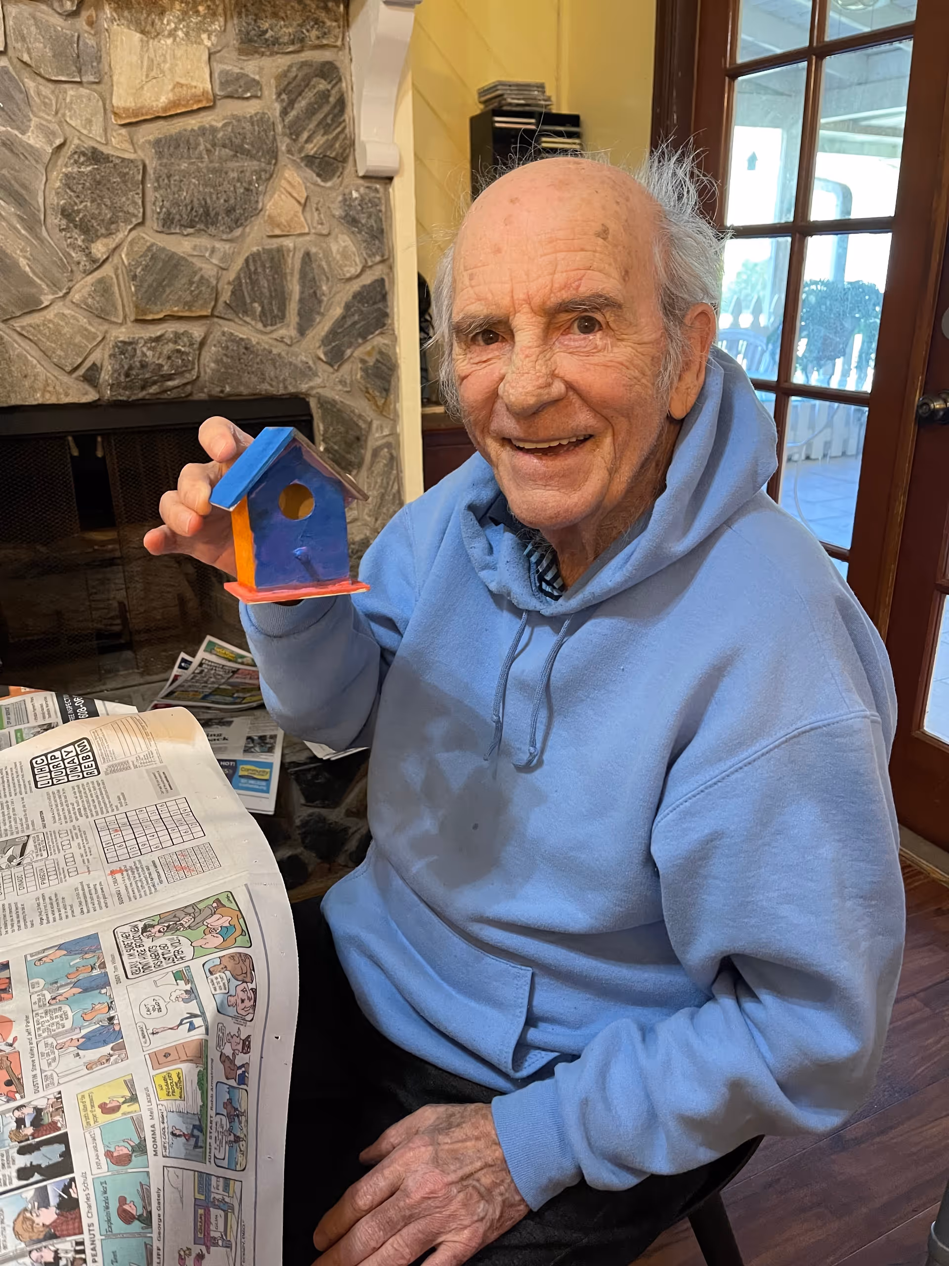 An elderly man wearing a light blue hoodie is sitting indoors near a stone fireplace and holding a small colorful birdhouse. He is smiling and there is a newspaper with comics on the table in front of him. Behind him, there is a glass door leading to an outdoor area.