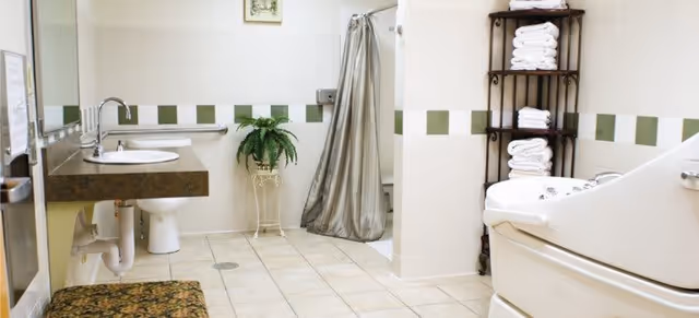 A spacious bathroom with beige tiled floor and white tiled walls featuring a green stripe. The room includes a wall-mounted sink with a mirror above it, a toilet, a shower area with a silver curtain, a potted plant on a stand, a cushioned bench, a shelf stocked with folded white towels, and a large white bathtub with jets.