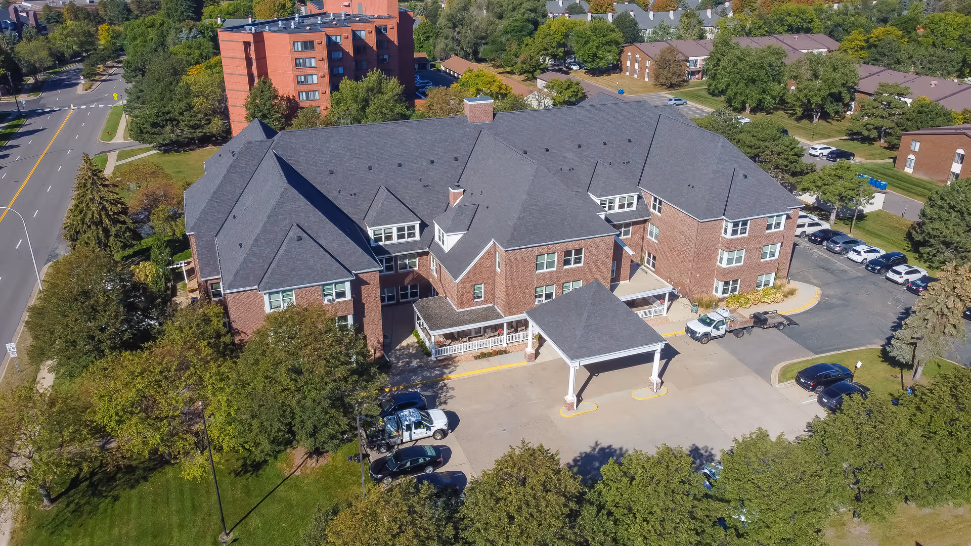 Aerial view of a large brick senior living facility with a dark gray roof, surrounded by trees and parking areas. The building has multiple wings and a covered entrance with a driveway. Nearby roads and other residential buildings are visible in the background.