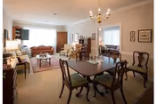 A spacious living and dining area in a retirement community featuring a wooden dining table with six chairs, a chandelier overhead, and a sitting area with sofas, armchairs, and a coffee table. The room is decorated with framed pictures on the walls and has large windows allowing natural light to fill the space.
