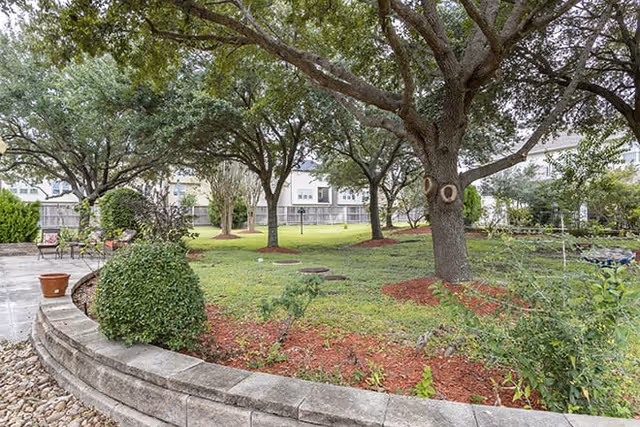 A landscaped outdoor garden area with large trees providing shade, green grass, and mulched planting beds. There is a curved stone retaining wall with some bushes and plants, and a paved patio area with chairs and a small table. Residential buildings are visible in the background.