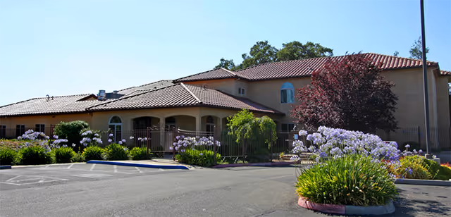Exterior view of a single-story building with a tiled roof and arched windows, surrounded by landscaped bushes and flowering plants under a clear blue sky.