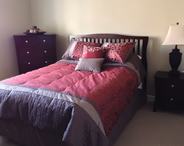 A neatly made bed with a dark wooden headboard, covered with a red and gray comforter and several decorative pillows. To the left of the bed is a dark wooden dresser with silver knobs and a decorative item on top. To the right of the bed is a matching dark wooden nightstand with a lamp featuring a white lampshade.