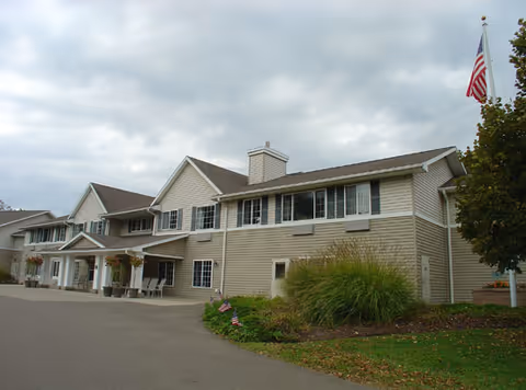 Two-story beige senior living facility building with a driveway, landscaping and an American flag under a cloudy sky.
