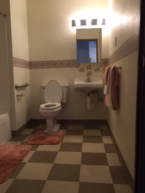 A bathroom with a toilet, a small wall-mounted sink with a mirror above it, and a towel rack holding pink and white towels. The floor has a checkered pattern with brown and beige tiles, and there are pink bath mats on the floor. The walls are light-colored with a decorative floral border around the middle.