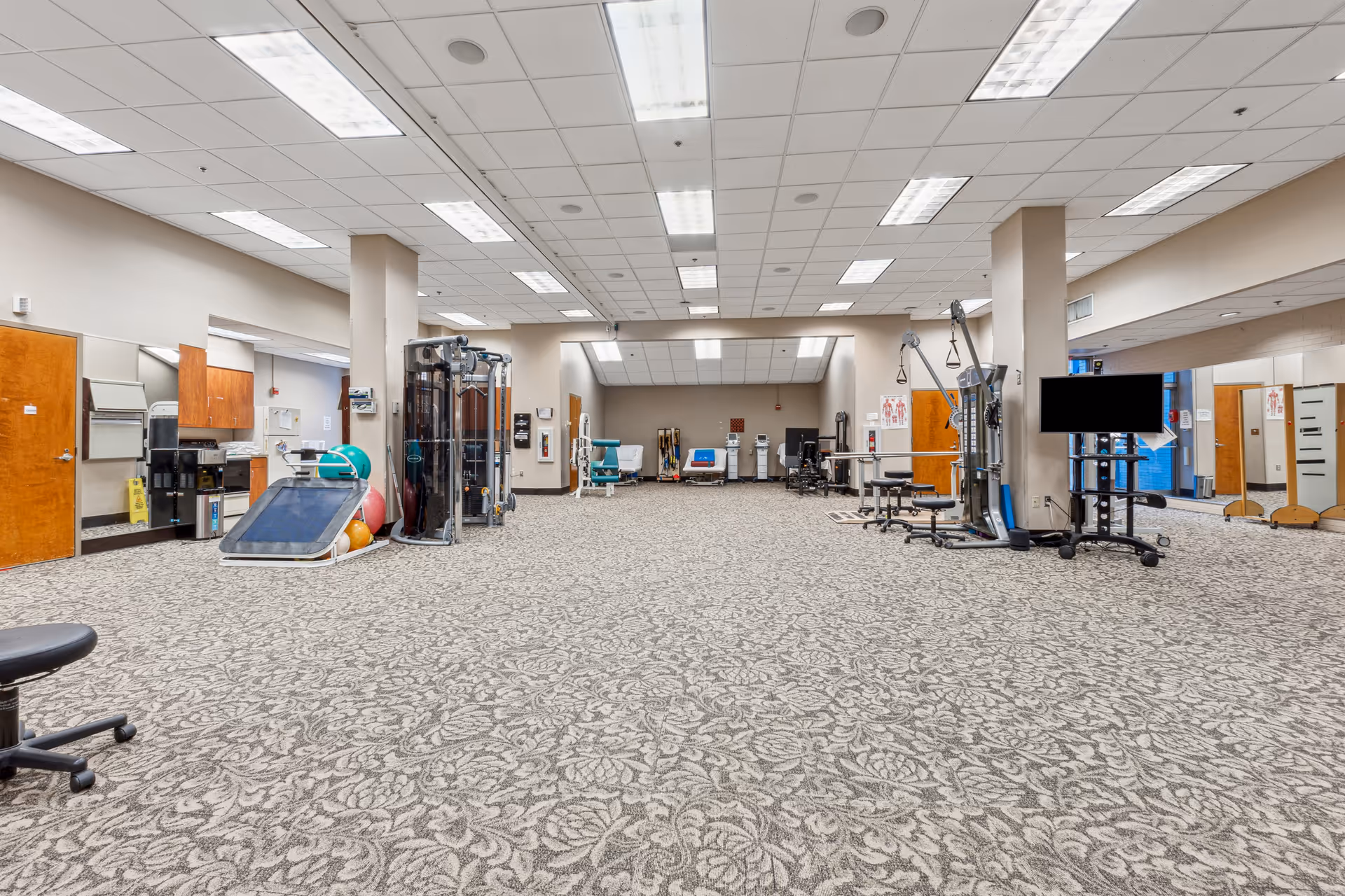 A large carpeted rehab/exercise room with various fitness machines, therapy equipment, and a TV.