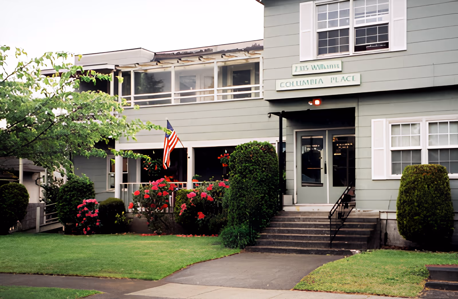 Exterior view of a two-story building with light green siding and white trim. There is a small staircase leading to double doors, an American flag displayed near the entrance, and well-maintained bushes and flowering plants in front of the building. A sign above the door reads 'Columbia Place'.