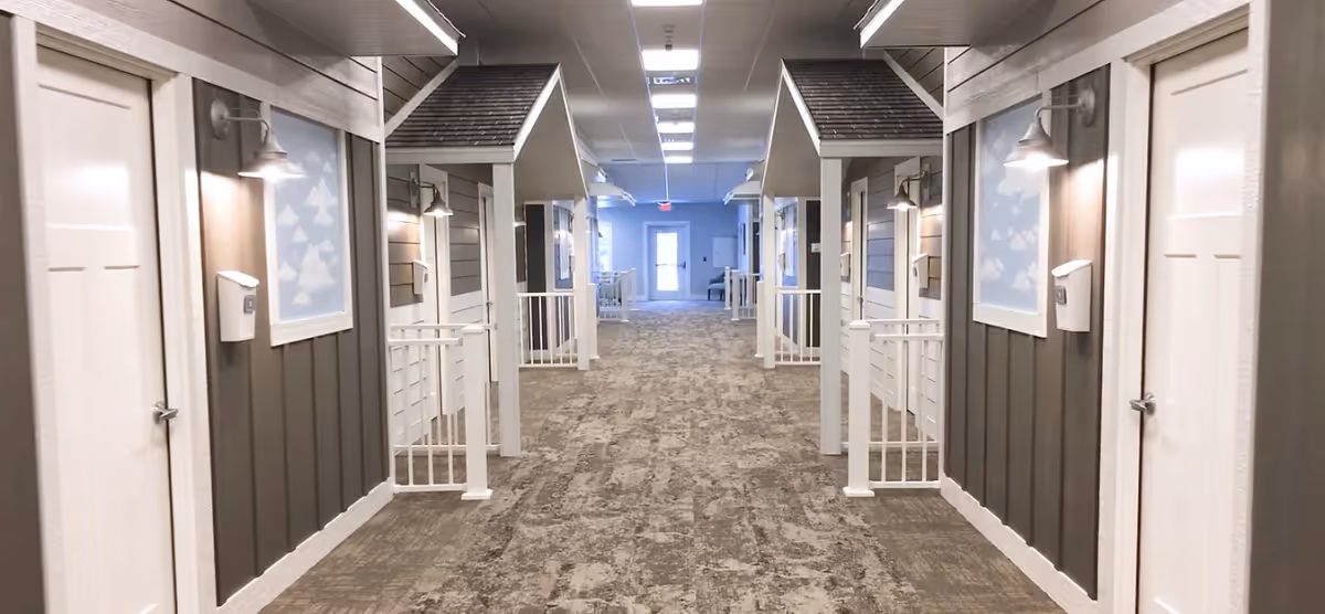 Well-lit interior hallway featuring mock porch facades and doors lining both sides leading to a distant exit.