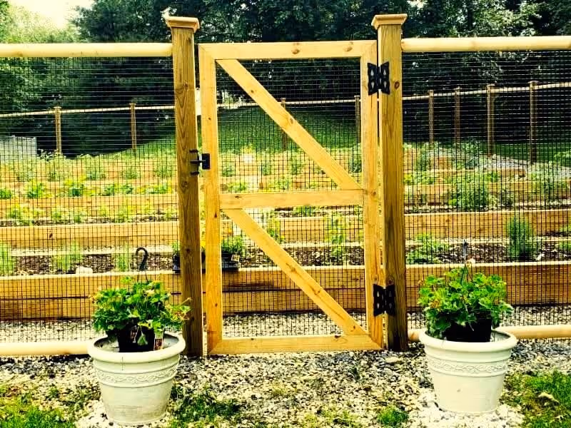 A wooden garden gate with wire mesh fencing on either side, enclosing a garden with raised planting beds. Two white flower pots with green plants are placed symmetrically in front of the gate on a gravel surface. Trees and greenery are visible in the background.