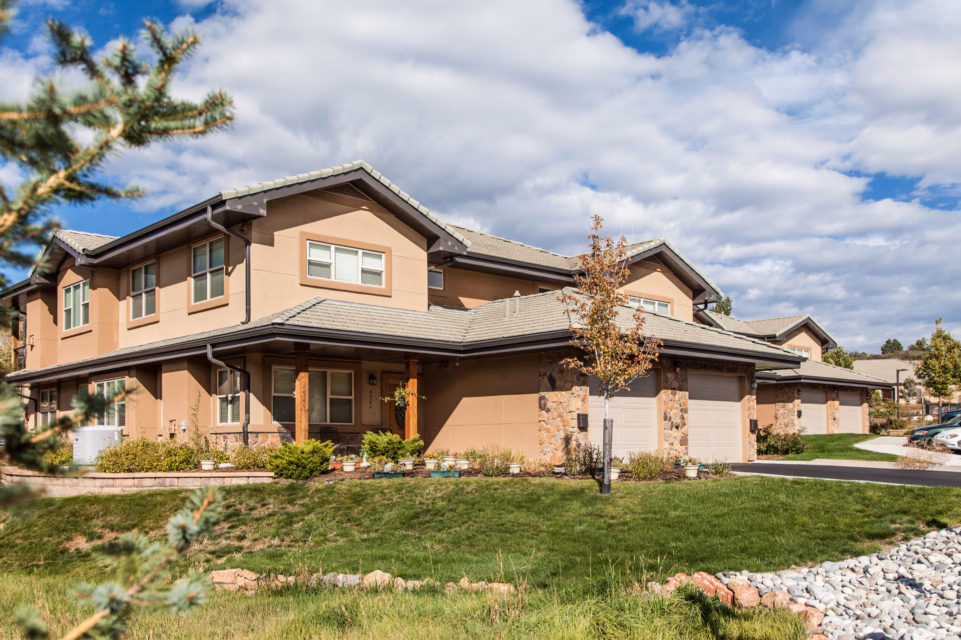 Exterior view of a two-story beige residential building with garages, landscaped lawn and cloudy sky.