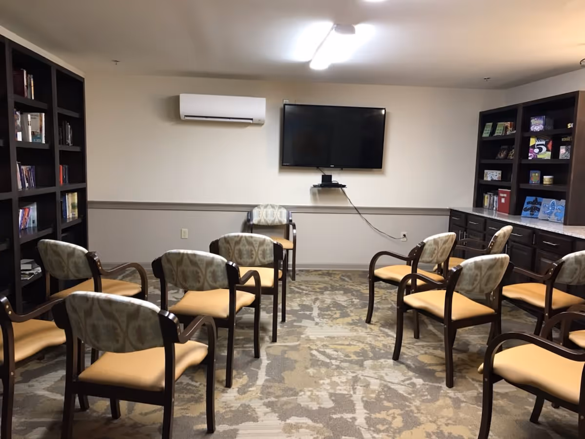 A small meeting or activity room with several chairs arranged in rows facing a wall-mounted flat screen TV. The room has bookshelves on both sides filled with books and decorative items, a carpeted floor with a patterned design, and a wall-mounted air conditioning unit above the TV.