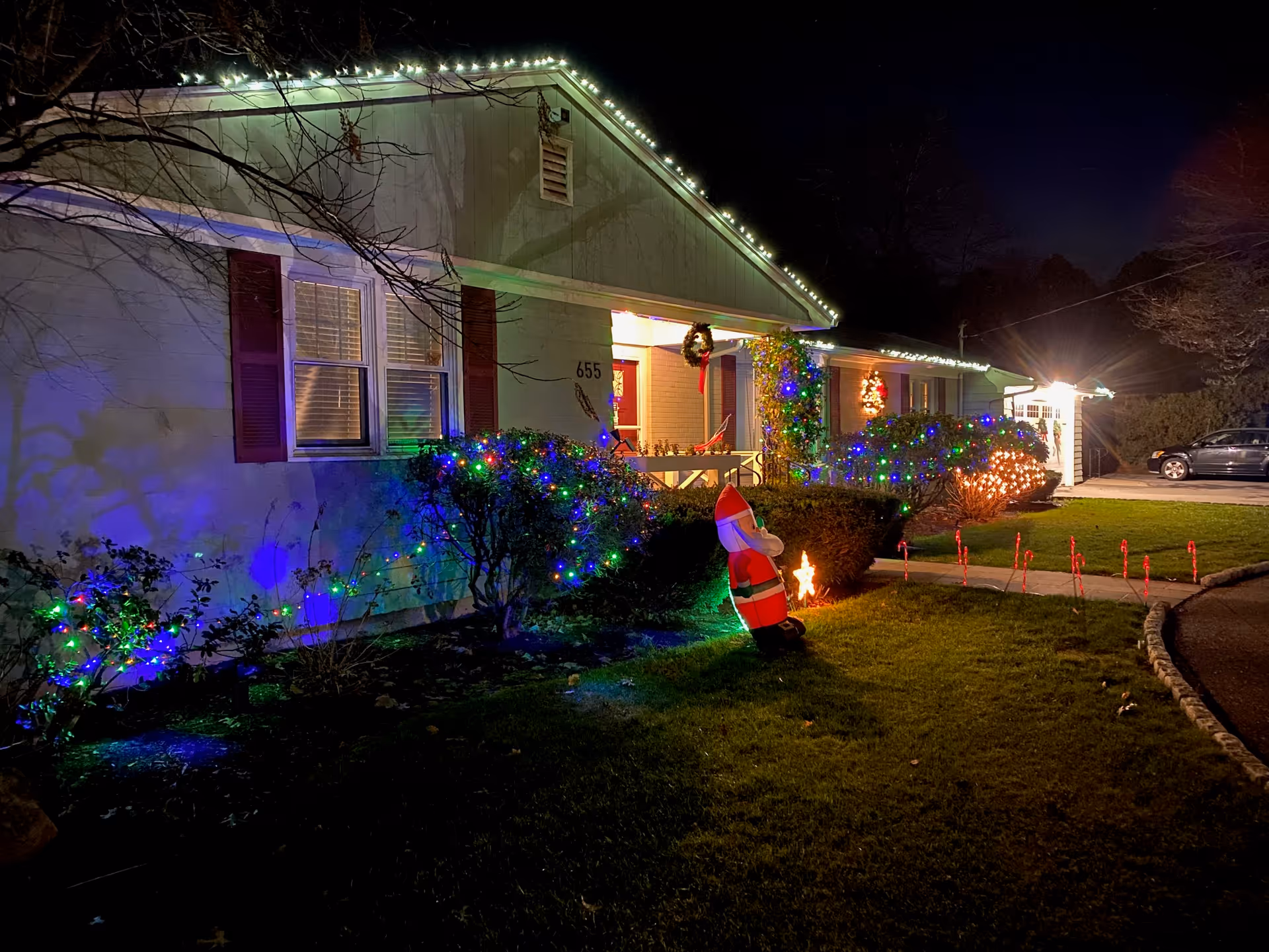 Front of a single-story house at night decorated with Christmas lights, lit bushes, and an inflatable Santa on the lawn.