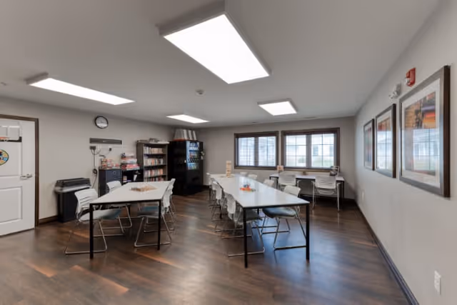 A well-lit common room with two long white tables surrounded by chairs, a vending machine, bookshelves, and windows with blinds letting in natural light. The room has wooden flooring, framed pictures on the wall, and overhead rectangular fluorescent lights.