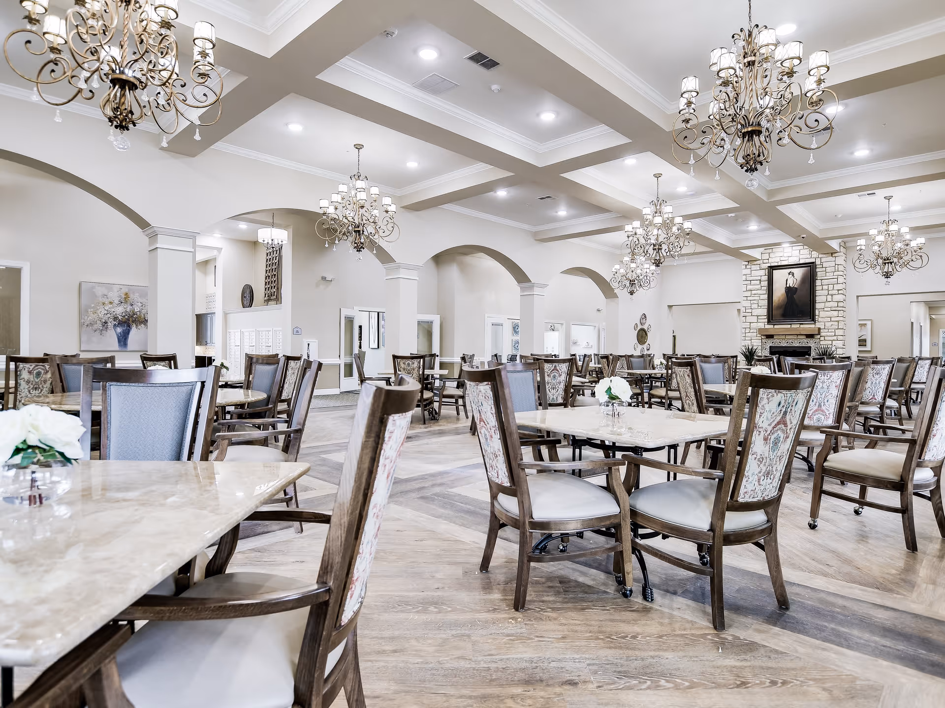 A spacious and elegant dining room with multiple tables and chairs arranged neatly. The room features high ceilings with decorative beams, several ornate chandeliers, a stone fireplace with a framed portrait above it, and light-colored walls with arched doorways. The floor is wooden, and small floral centerpieces are placed on the tables.