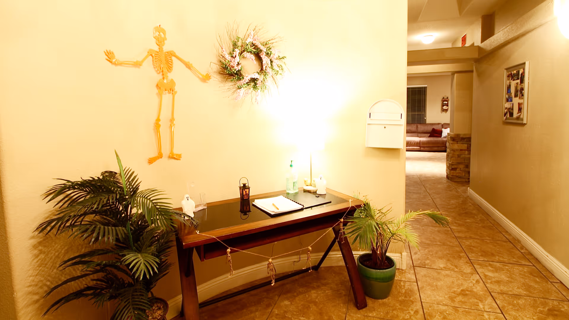 A hallway with beige walls and tiled floor featuring a small wooden table with a glass top. On the table are a lamp, a notebook, a hand sanitizer bottle, and small decorative items. Above the table on the wall is a decorative wreath and a skeleton decoration. There are two potted plants on either side of the table. The hallway leads to a room with a brown couch and a window with blinds.