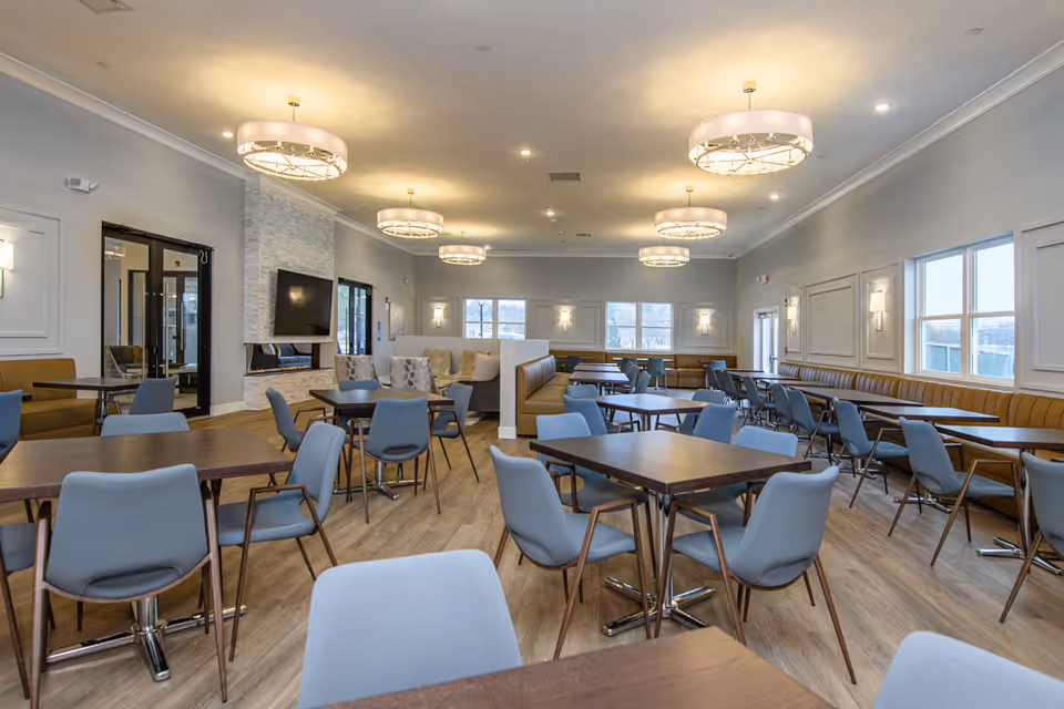 A spacious and well-lit dining room with multiple wooden tables and blue cushioned chairs arranged neatly. The room features modern circular ceiling lights, large windows allowing natural light, a fireplace with a mounted TV above it, and a combination of booth and chair seating along the walls.