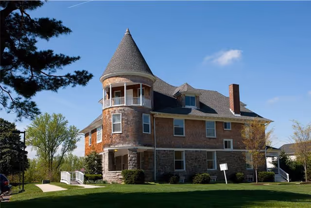 A large two-story building with a conical turret, shingle siding, and stone foundation surrounded by green grass and trees under a clear blue sky.