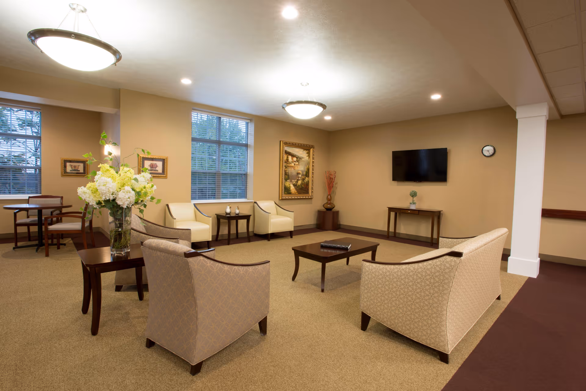 A well-lit living room area in a senior living facility with beige walls and carpet. The room features comfortable armchairs and sofas arranged around a wooden coffee table, a flat-screen TV mounted on the wall, framed artwork, and a vase with flowers on a side table. Large windows let in natural light.