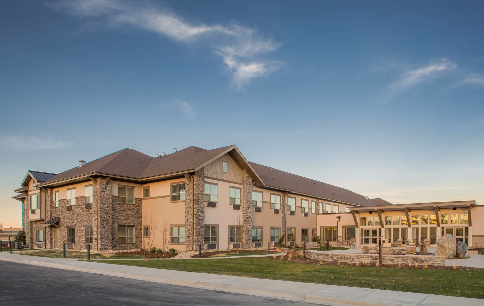 Two-story senior living facility building with stone accents, many windows and an outdoor seating area under a blue sky.