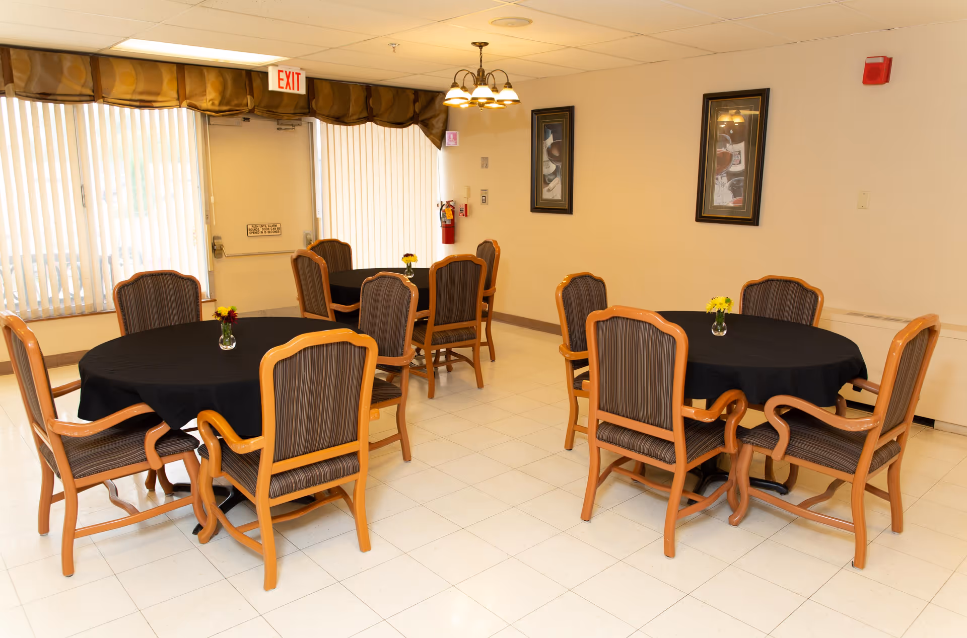 Interior view of a dining room with two round tables covered with black tablecloths, each surrounded by six wooden chairs with striped cushions. Small flower vases are placed in the center of each table. The room has tiled flooring, beige walls with two framed pictures, vertical blinds covering the windows, and an exit door with an illuminated exit sign above it.