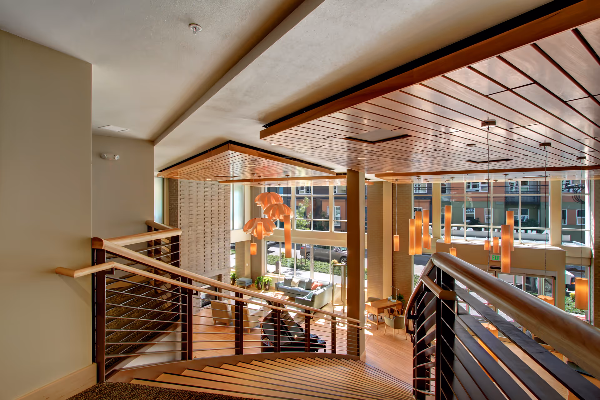 Spacious two-story interior lobby seen from a curved staircase overlooking seating areas, pendant lights, and large windows.