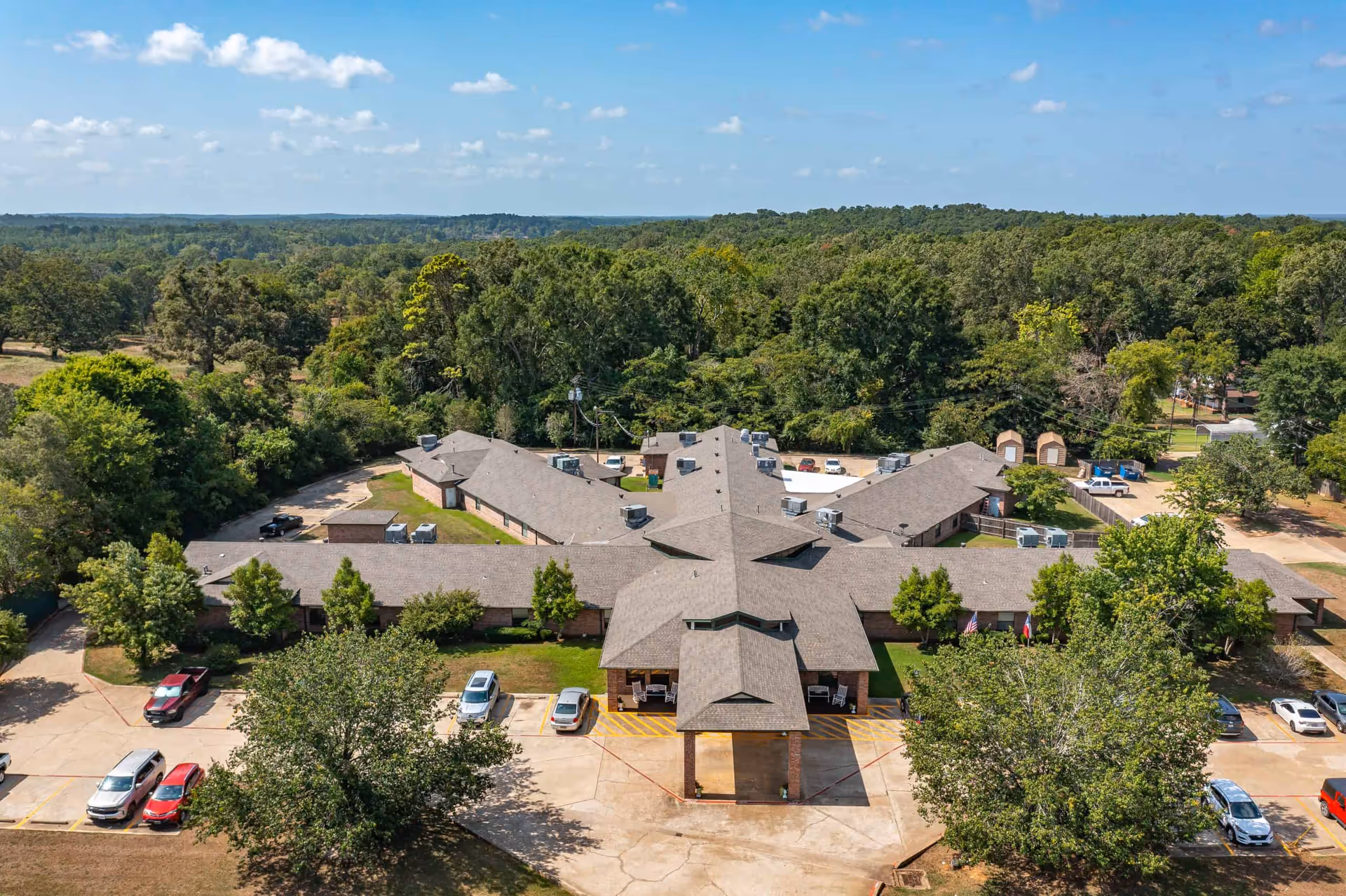 Aerial view of Summit Nursing & Rehab of San Augustine, a single-story building with a star-shaped roof surrounded by trees and greenery. Several cars are parked in the parking lot in front of the building under a partly cloudy sky.