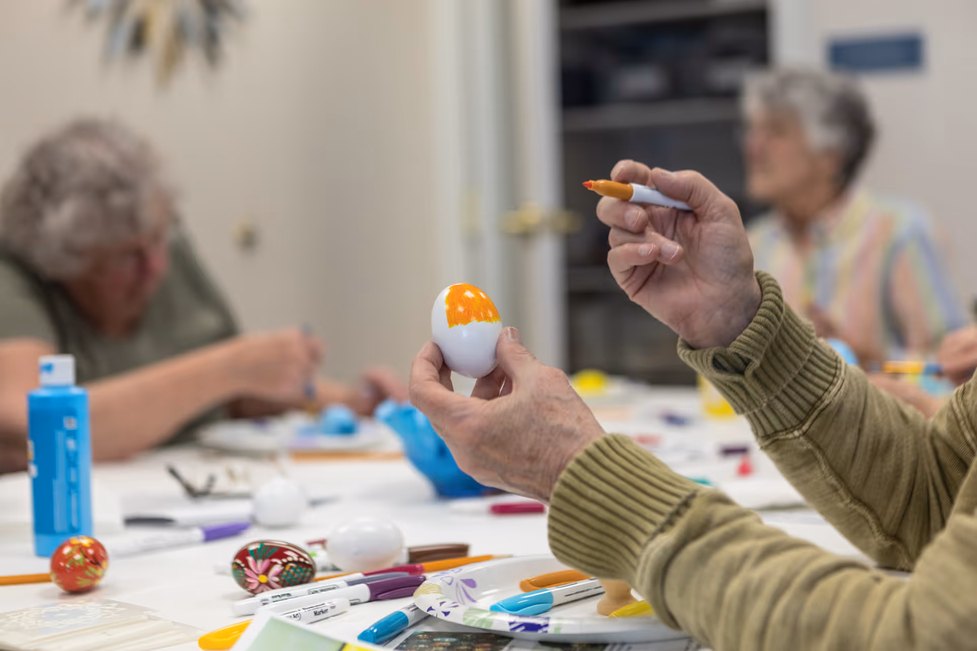 Two elderly individuals sitting at a table engaged in painting and decorating eggs with various colored markers and paints, focusing on one person's hands holding a white egg partially painted orange.